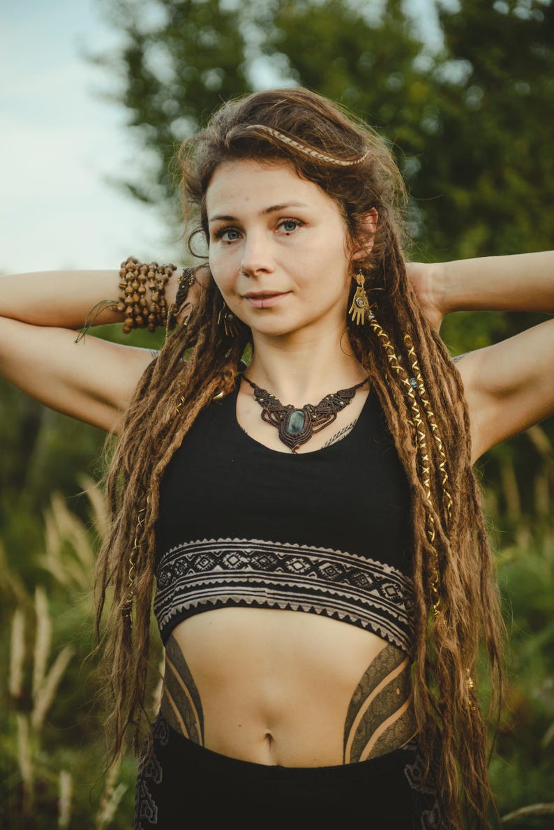 May include: A black crop top featuring a wide band with a geometric pattern, paired with a macrame necklace containing a central dark stone. The jewelry includes dangling hand-shaped earrings and a beaded bracelet on the wrist.