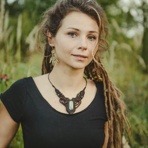 May include: A dark brown macrame necklace featuring a central oval-shaped light green stone, accented with small metallic beads. The necklace is paired with gold-toned earrings shaped like open palms, each containing a small circular green stone inlay.