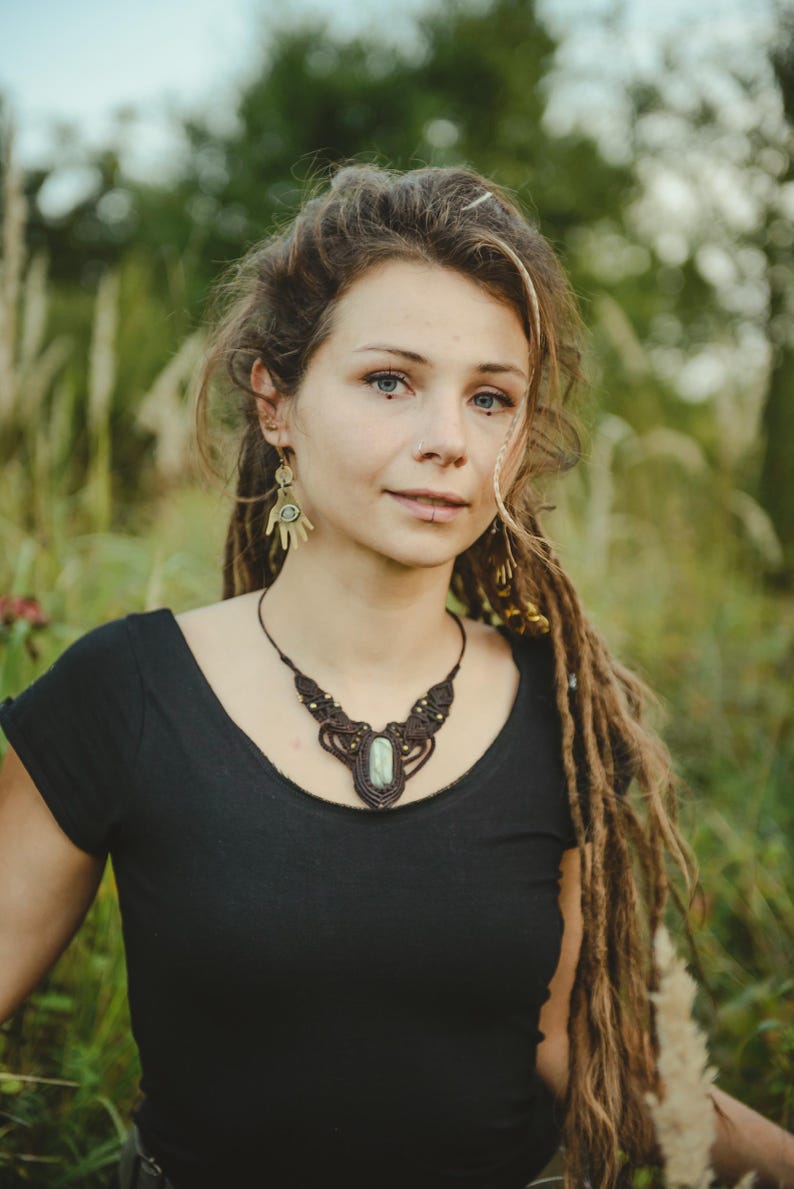 May include: A dark brown macrame necklace featuring a central oval-shaped light green stone, accented with small metallic beads. The necklace is paired with gold-toned earrings shaped like open palms, each containing a small circular green stone inlay.