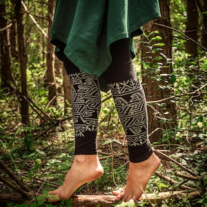 Op de afbeelding: Zwarte leggings met een wit tribaal patroon. De persoon draagt een groene tuniek en staat blootsvoets in een bos, omringd door bomen en bladeren. De leggings zijn het middelpunt van de afbeelding.