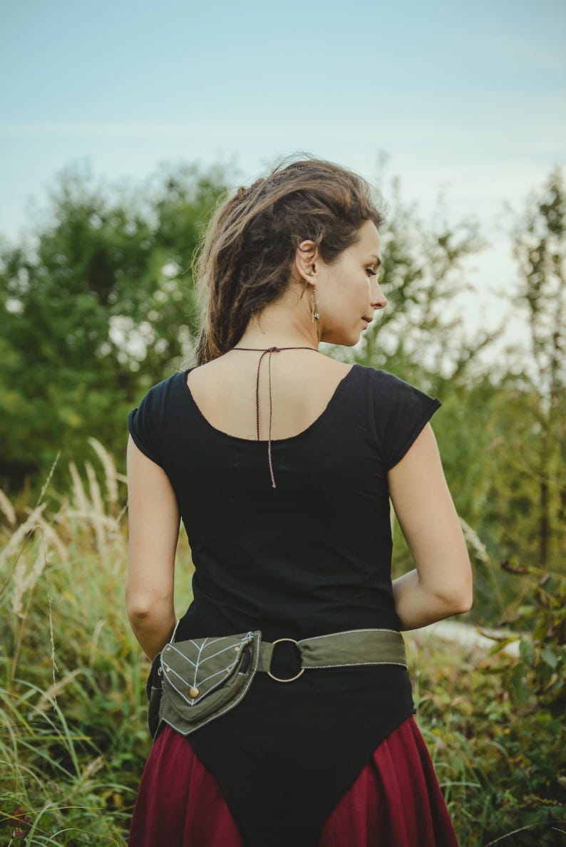 May include: A black short-sleeved top with a scoop neckline and a thin tie at the back of the neck. An olive green belt features a leaf-shaped pouch with white stitching, a metallic snap button, and a circular metal ring attachment at the waist.