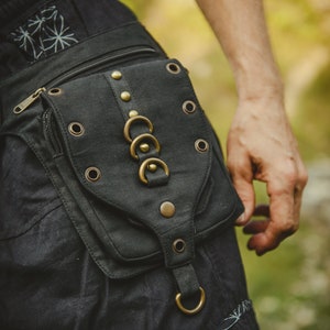 Könnte beinhalten: Ein schwarzer Canvas-Gürtel mit einer großen Tasche und dekorativen Metallringen. Die Tasche hat einen Reißverschluss und mehrere Nieten aus Metall.
