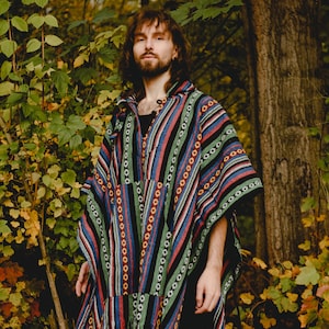 May include: A person wearing a colourful striped poncho with a hood. The poncho has a black background with red, blue, green and yellow stripes. The person is standing in a woodland setting with autumn foliage.