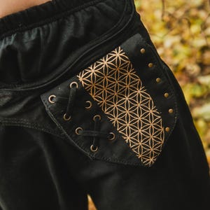 Può includere: Pantaloni neri con una tasca decorativa con un motivo geometrico dorato. La tasca ha occhielli e un dettaglio di allacciatura. Rivetti dorati bordano il bordo della tasca. I pantaloni hanno una vita con coulisse.