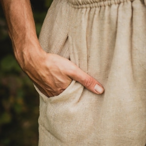 May include: Close-up of beige linen pants with an elastic waistband and a side pocket. The fabric has a textured appearance. The hand is placed inside the pocket.