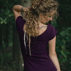 May include: A dark purple short-sleeved t-shirt with a wide scoop neckline, featuring a small rectangular label on the side seam. The garment is worn with a patterned skirt in shades of mustard yellow, brown, and white with a floral print.