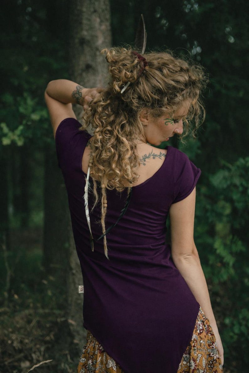 May include: A dark purple short-sleeved t-shirt with a wide scoop neckline, featuring a small rectangular label on the side seam. The garment is worn with a patterned skirt in shades of mustard yellow, brown, and white with a floral print.