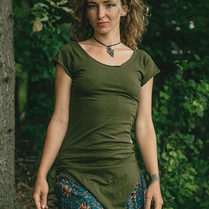 May include: An olive green short-sleeved tunic with a scoop neckline and a pointed hemline, paired with a teal skirt featuring a floral pattern in shades of orange, brown, and cream. The tunic has a fitted silhouette and raw-edge sleeve details.