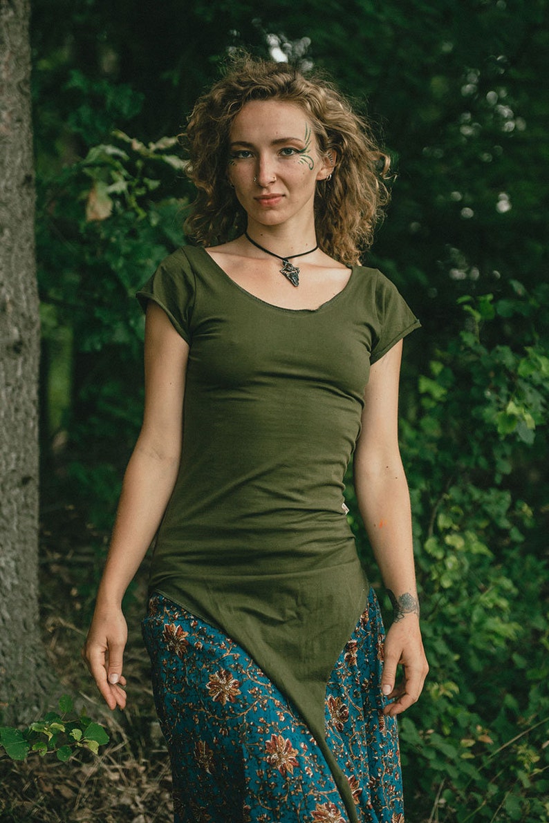 May include: An olive green short-sleeved tunic with a scoop neckline and a pointed hemline, paired with a teal skirt featuring a floral pattern in shades of orange, brown, and cream. The tunic has a fitted silhouette and raw-edge sleeve details.