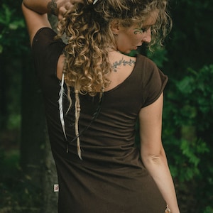May include: A dark brown short-sleeved top with a scoop neckline and a small rectangular tag on the lower side seam. The garment is fitted, featuring a plain back design and short sleeves with folded cuffs, worn with a patterned skirt.