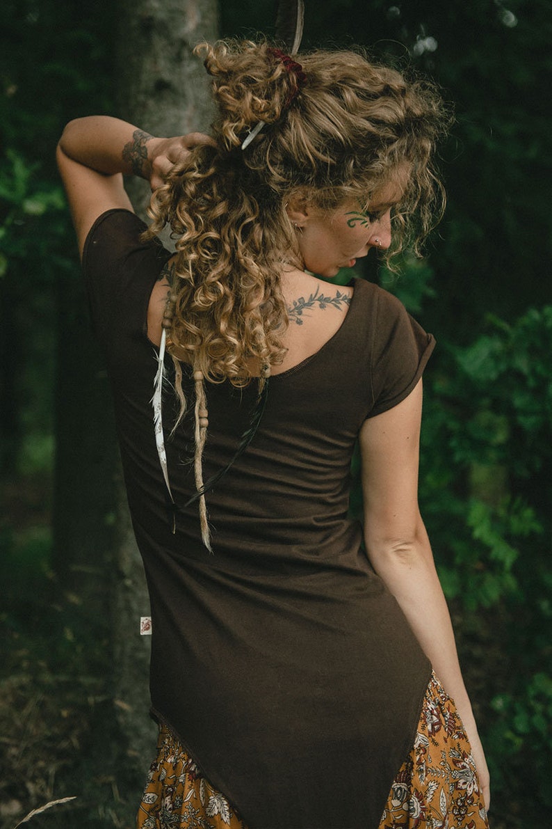 May include: A dark brown short-sleeved top with a scoop neckline and a small rectangular tag on the lower side seam. The garment is fitted, featuring a plain back design and short sleeves with folded cuffs, worn with a patterned skirt.