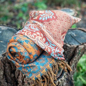 May include: A rolled scarf featuring a vibrant orange and blue floral pattern with a contrasting red and blue geometric border, finished with fringed edges. The text FREE SILKY GIFT BAG FOR EVERY PURCHASE is displayed in white at the top and bottom.