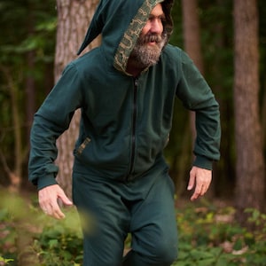 Peut inclure: Combinaison à capuche vert foncé avec capuche pointue et bordure décorative. La combinaison a une fermeture éclair intégrale et un pantalon assorti. La personne est pieds nus dans une forêt. L'ensemble évoque un elfe des bois.