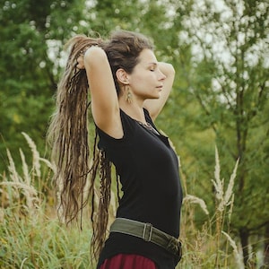 May include: A black short-sleeved top with a high-low hemline, paired with a dark red skirt and an olive green canvas belt featuring a metal buckle. The outfit is worn with long, textured brown dreadlocks and dangling earrings.
