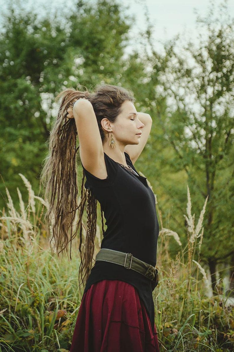 May include: A black short-sleeved top with a high-low hemline, paired with a dark red skirt and an olive green canvas belt featuring a metal buckle. The outfit is worn with long, textured brown dreadlocks and dangling earrings.