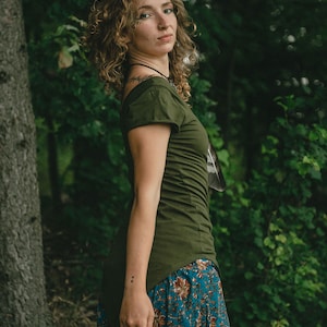May include: An olive green short-sleeved top with a high-low hemline paired with a teal blue skirt featuring a repeating floral pattern in shades of orange, brown, and cream. The top has a wide neckline and a fitted silhouette, while the skirt is pleated.