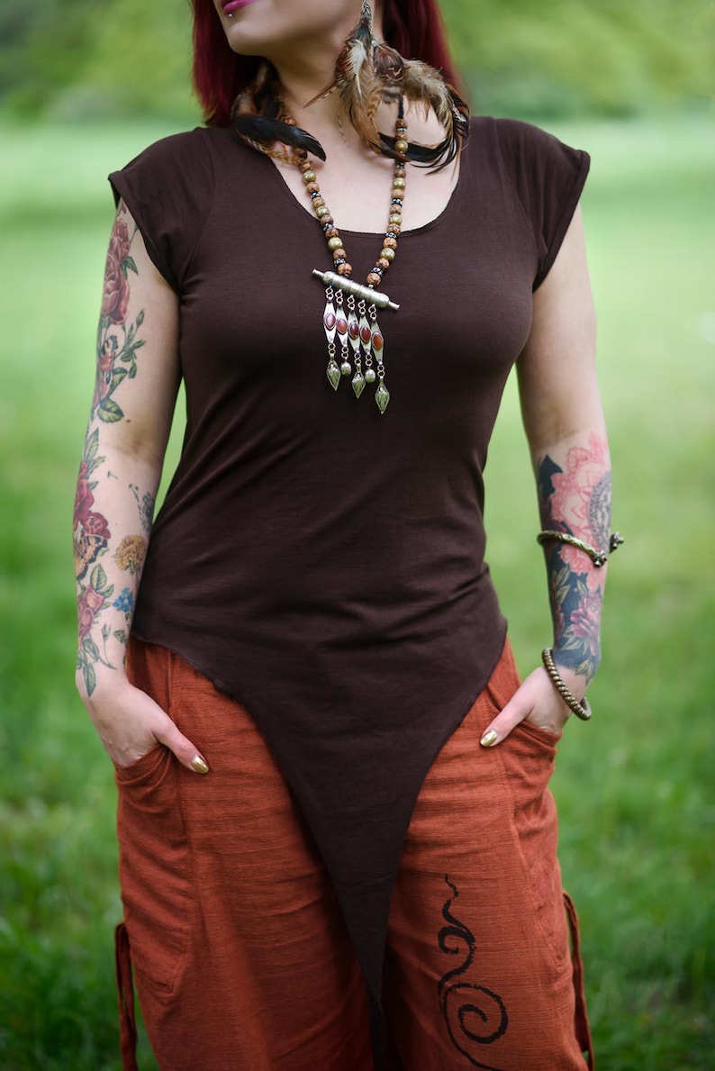 May include: A dark brown short-sleeved top with a scoop neckline and a long, pointed asymmetrical hemline. The top is paired with loose-fitting burnt orange trousers featuring side pockets and a black spiral graphic on the lower left leg.