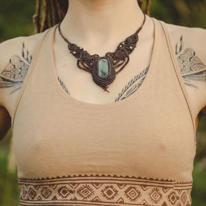 May include: A beige crop top featuring a dark brown geometric pattern along the hem, paired with a dark brown macrame necklace containing a central grey stone and small gold-toned beads, and brass-toned hand-shaped earrings with eye details.