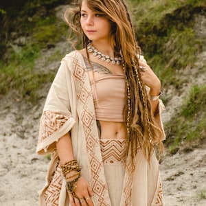 Può includere: Una donna indossa un completo beige con motivi geometrici marroni. L'outfit comprende una giacca lunga, un top corto e una gonna. Indossa anche una collana di conchiglie, braccialetti e anelli. I suoi lunghi capelli sono mossi con dreadlocks.