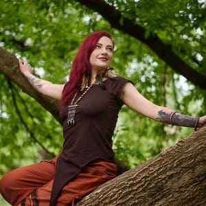 May include: A dark brown short-sleeved top with an asymmetrical pointed hemline, paired with loose-fitting rust-colored trousers featuring dark circular patterns. The outfit is accessorized with a long beaded necklace with metallic accents.