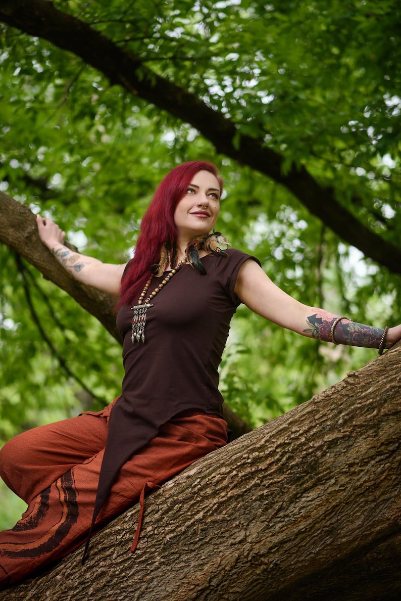 May include: A dark brown short-sleeved top with an asymmetrical pointed hemline, paired with loose-fitting rust-colored trousers featuring dark circular patterns. The outfit is accessorized with a long beaded necklace with metallic accents.