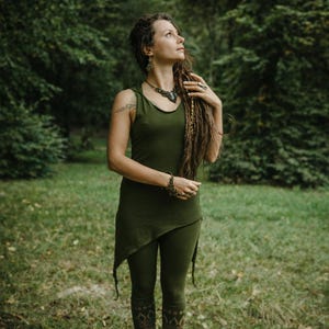 May include: An olive green outfit comprising a tank top, leggings, and boots. The tank top has an asymmetrical hemline. The leggings feature a decorative pattern on the lower legs. The person is wearing jewellery, including a necklace and bracelets, and has long dreadlocks.
