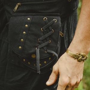 Może przedstawiać: Czarne spodnie z płótna z dużą, ozdobną kieszenią z sznurowaniem i złotymi akcentami. Kieszeń ma zamek błyskawiczny i jest zabezpieczona nitami.