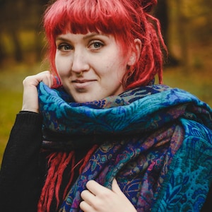 May include: A large, rectangular scarf featuring a complex, multi-coloured paisley and floral pattern in shades of deep blue, teal, purple, and magenta. The fabric has a soft, woven texture and is wrapped around the neck and shoulders over a black jumper.