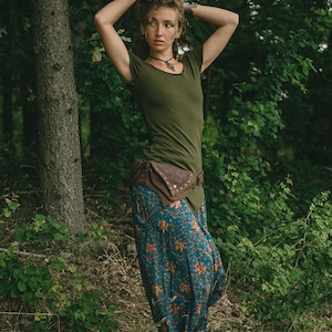May include: An olive green short-sleeved t-shirt paired with loose-fitting blue trousers featuring a repeating orange and white floral pattern. A brown leather belt with an attached pouch sits at the waist, and a dark pendant hangs from a cord.