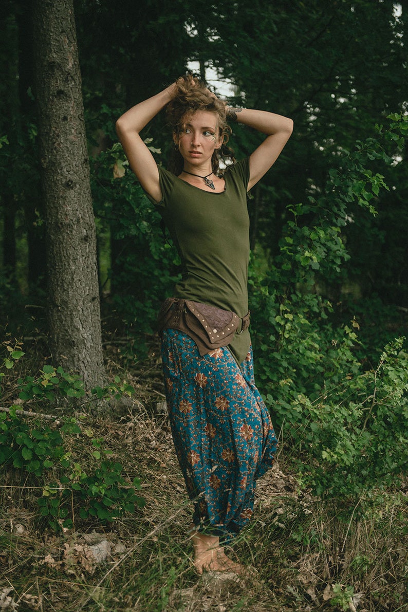 May include: An olive green short-sleeved t-shirt paired with loose-fitting blue trousers featuring a repeating orange and white floral pattern. A brown leather belt with an attached pouch sits at the waist, and a dark pendant hangs from a cord.
