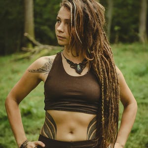 May include: A dark brown crop top with a scoop neckline and matching dark brown leggings. The outfit is paired with a dark macrame necklace featuring a central stone pendant and a matching macrame hand accessory with a small embedded stone.