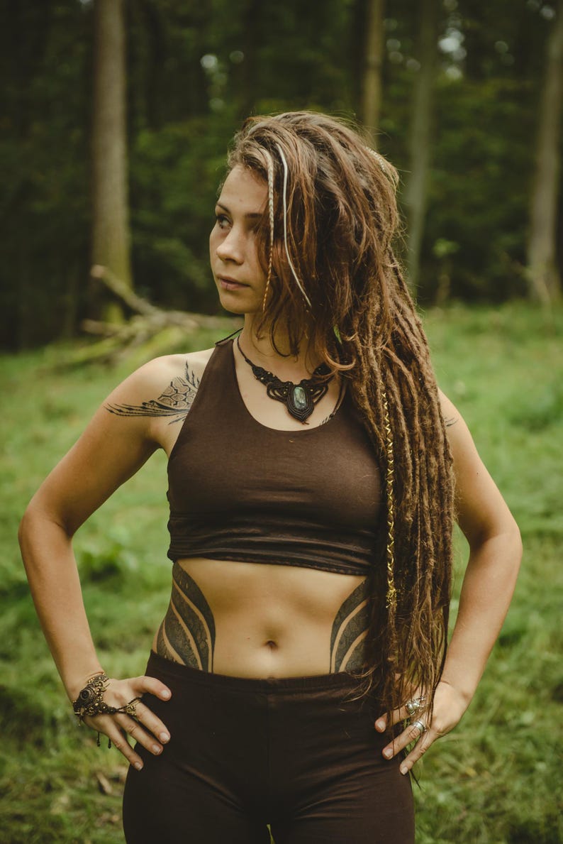 May include: A dark brown crop top with a scoop neckline and matching dark brown leggings. The outfit is paired with a dark macrame necklace featuring a central stone pendant and a matching macrame hand accessory with a small embedded stone.