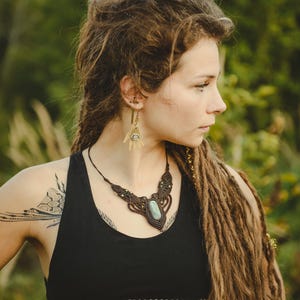 May include: A macrame necklace featuring a central oval green stone set in dark brown cord with intricate knotting and small metallic accents. The jewelry is paired with brass-toned earrings shaped like open palms with a central eye motif.