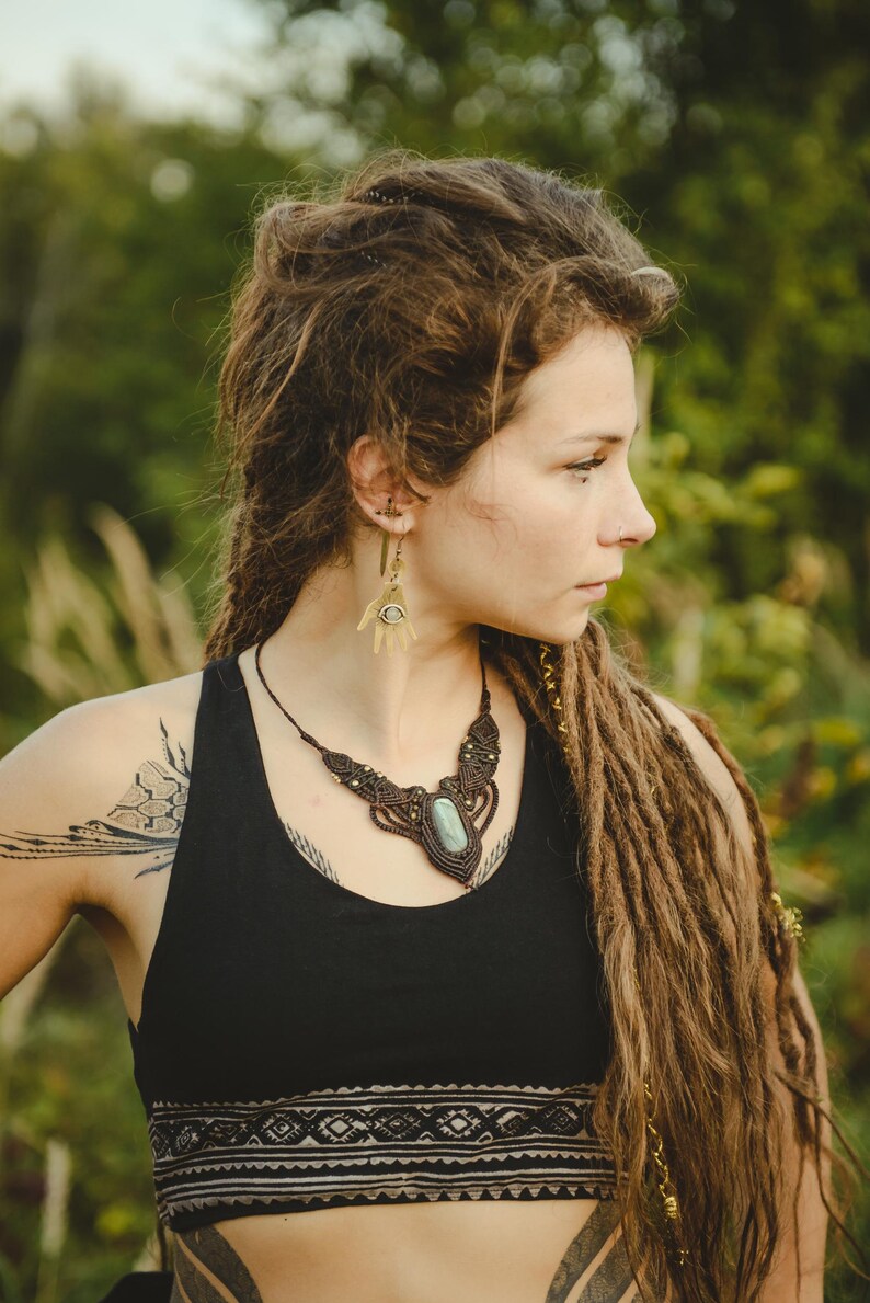 May include: A macrame necklace featuring a central oval green stone set in dark brown cord with intricate knotting and small metallic accents. The jewelry is paired with brass-toned earrings shaped like open palms with a central eye motif.