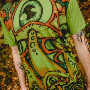 May include: Green t-shirt featuring a psychedelic print of a mushroom with an eye. The shirt has the word "mirror" printed on the shoulder.