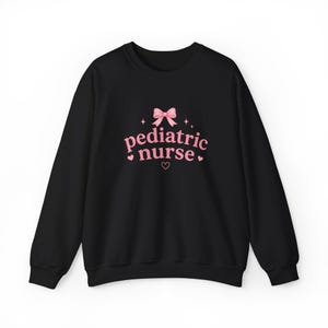Op de afbeelding: Een zwart sweatshirt met lange mouwen en een roze afbeelding op de borst. Het ontwerp bevat een roze strik aan de bovenkant, de tekst "pediatric nurse" in een roze schreeflettertype en kleine roze hartjes en sterretjes rondom de tekst.