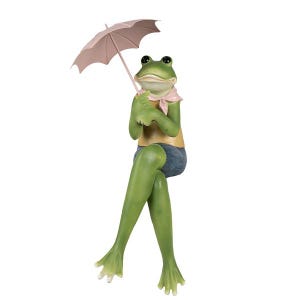 Könnte beinhalten: Eine grüne Froschfigur mit einer rosa Schleife und einem rosa Regenschirm. Der Frosch sitzt mit gekreuzten Beinen und trägt ein gelbes Hemd und eine blaue Shorts.