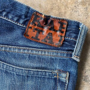 Puede incluir: Primer plano de unos vaqueros de mezclilla azules con un parche de cuero marr&oacute;n. El parche presenta la palabra "TA" en un dise&ntilde;o estilizado. Los vaqueros tienen un dise&ntilde;o cl&aacute;sico de cinco bolsillos y est&aacute;n hechos de tela de mezclilla duradera.