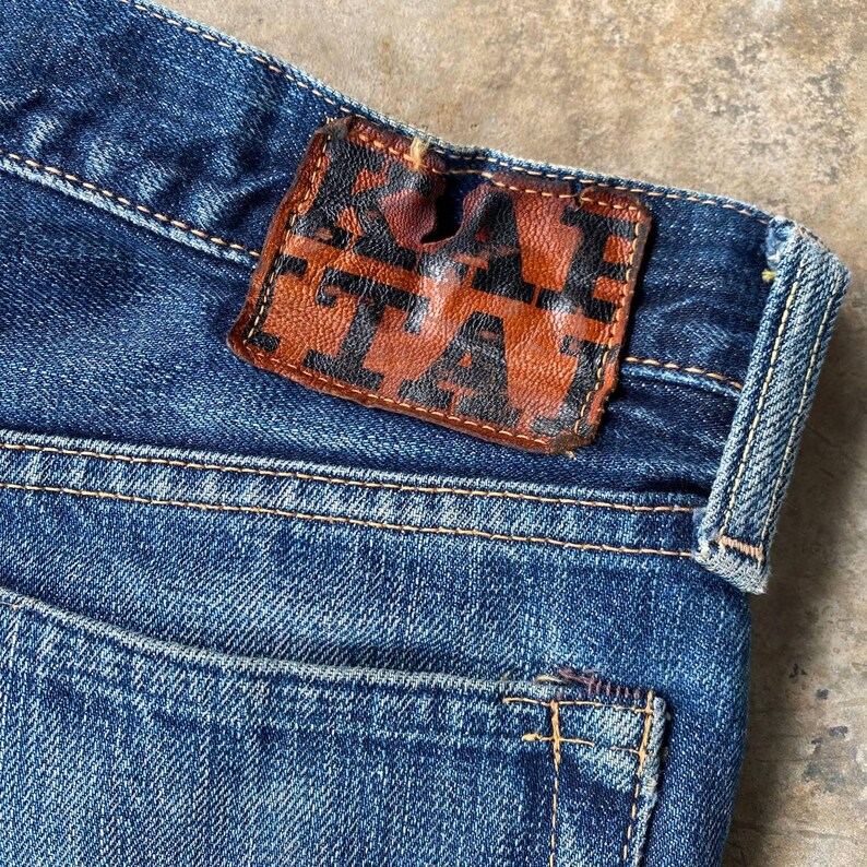 Puede incluir: Primer plano de unos vaqueros de mezclilla azules con un parche de cuero marr&oacute;n. El parche presenta la palabra "TA" en un dise&ntilde;o estilizado. Los vaqueros tienen un dise&ntilde;o cl&aacute;sico de cinco bolsillos y est&aacute;n hechos de tela de mezclilla duradera.
