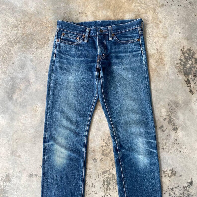 Puede incluir: Vaqueros de mezclilla azul descolorido con un dise&ntilde;o cl&aacute;sico de cinco bolsillos. Los vaqueros tienen bragueta con botones y corte recto. La mezclilla muestra signos de desgaste y decoloraci&oacute;n, d&aacute;ndole un aspecto vintage.