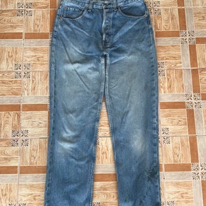 May include: Light blue denim jeans with a button fly and a classic five-pocket design. The jeans have a faded wash and a straight leg cut. The jeans are laid flat on a tiled surface.