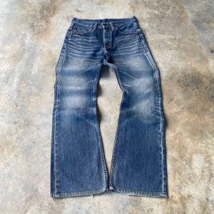 Puede incluir: Vaqueros azules de mezclilla con diseño de pernera acampanada. Los vaqueros tienen bragueta con botones, estilo clásico de cinco bolsillos y muestran signos de desgaste con zonas descoloridas. Los vaqueros están planos sobre una superficie de hormigón.