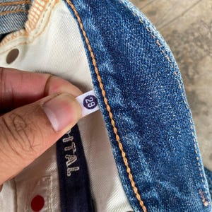 Puede incluir: Primer plano de unos vaqueros azules con una etiqueta de talla 29. Los vaqueros tienen un dise&ntilde;o cl&aacute;sico de cinco bolsillos, con costuras visibles y un lavado claro. La etiqueta es blanca con el n&uacute;mero 29 en un c&iacute;rculo negro.