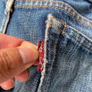 Puede incluir: Primer plano de unos vaqueros de mezclilla azules con una etiqueta roja. La etiqueta tiene texto blanco y est&aacute; cosida en la costura del bolsillo de los vaqueros. La mezclilla tiene un aspecto desgastado con costuras visibles.