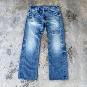 May include: A pair of blue denim jeans featuring a faded wash, whiskering at the hips, and a rectangular patch detail on the left thigh. The jeans have a classic five-pocket construction with a button fly, belt loops, and straight leg openings.