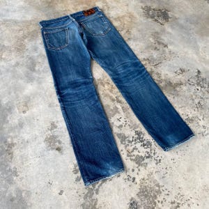 Puede incluir: Vaqueros de mezclilla azul descolorido con un dise&ntilde;o cl&aacute;sico de cinco bolsillos. Los vaqueros muestran desgaste, con decoloraci&oacute;n y desgaste visibles. Un parche de cuero marr&oacute;n es visible en la cintura trasera. Los vaqueros est&aacute;n planos.