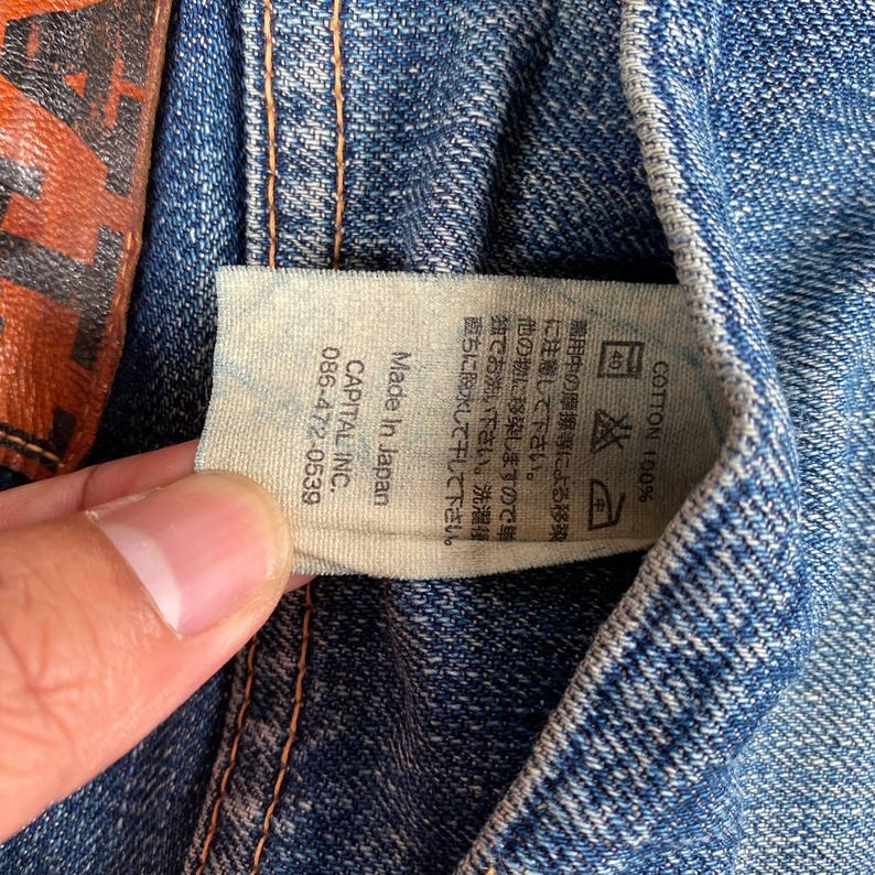 Puede incluir: Primer plano de una prenda de mezclilla azul con una etiqueta blanca. La etiqueta dice "Made in Japan" y "CAPITAL INC. 086-472-0539". La etiqueta tambi&eacute;n incluye instrucciones de lavado en japon&eacute;s y el texto "COTTON 100%."