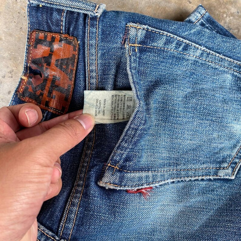 Puede incluir: Primer plano de unos vaqueros de mezclilla azules con un parche de cuero desgastado y una peque&ntilde;a etiqueta. El parche tiene un dise&ntilde;o marr&oacute;n y negro. La etiqueta tiene texto en japon&eacute;s e ingl&eacute;s. Los vaqueros tienen un dise&ntilde;o cl&aacute;sico de cinco bolsillos.