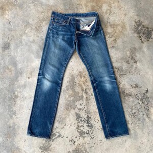 Puede incluir: Un par de vaqueros de mezclilla azul con bragueta de botones, colocados sobre una superficie de hormig&oacute;n. Los vaqueros tienen un dise&ntilde;o cl&aacute;sico de cinco bolsillos y muestran signos de desgaste, con zonas descoloridas y sutiles.