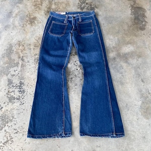 Puede incluir: Vaqueros de mezclilla azul oscuro con diseño de pernera acampanada. Los vaqueros tienen dos bolsillos delanteros y dos traseros. Las costuras son de color beige en contraste. Los vaqueros están colocados sobre una superficie de hormigón.